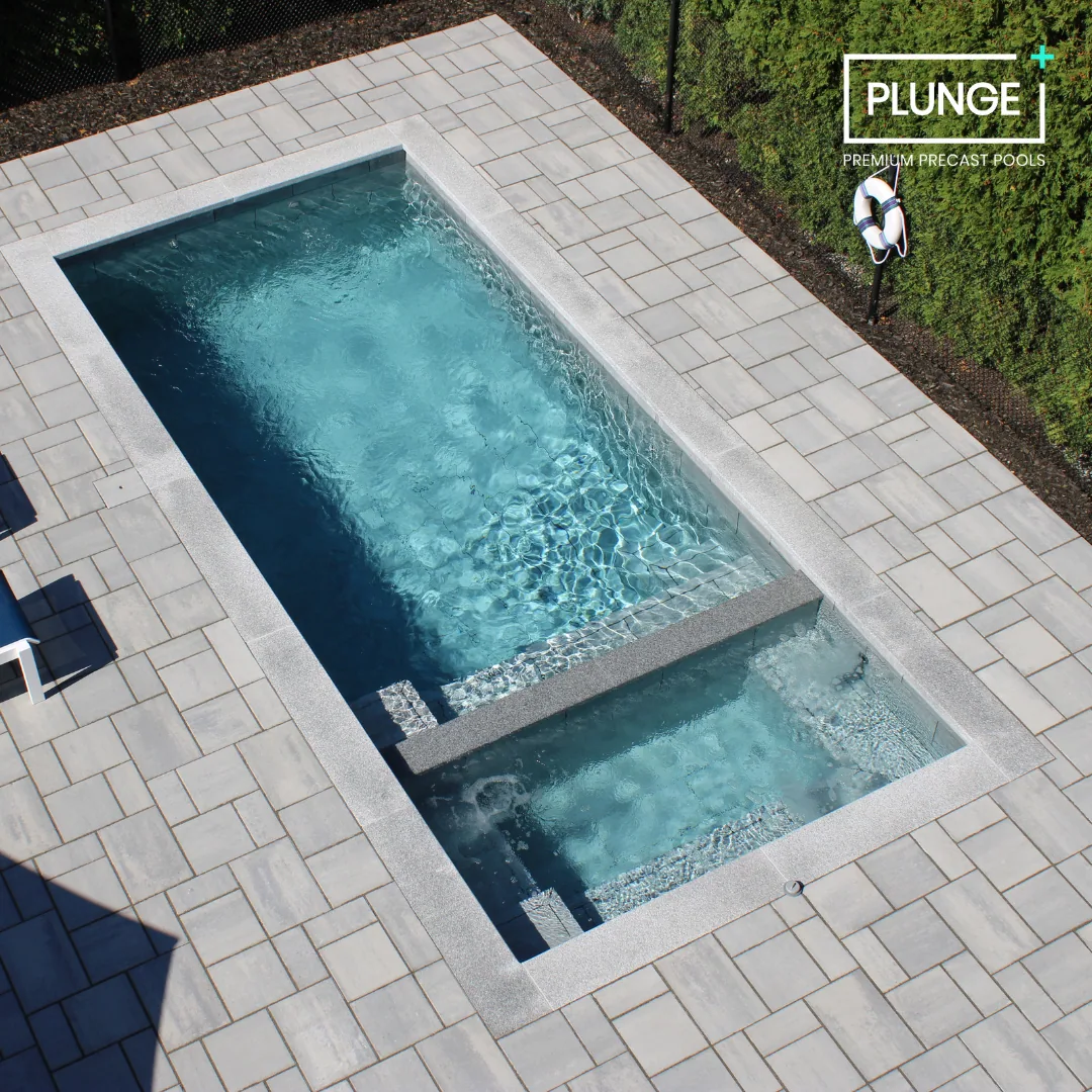 An outdoor rectangular swimming pool with a hot tub attached, surrounded by light-colored paving stones and backed by green bushes.