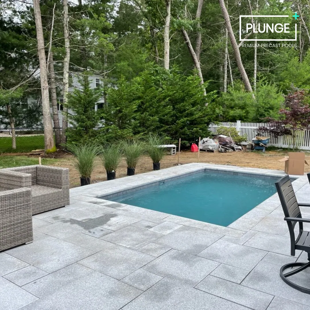 Backyard with a small rectangular pool, outdoor furniture, potted grasses, tall trees, and a white fence.