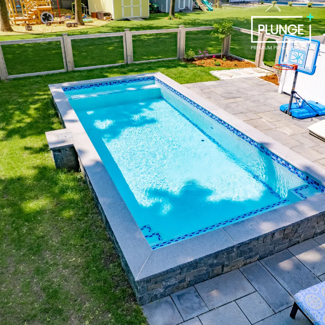 A backyard with a rectangular in-ground swimming pool with blue tiles, surrounded by a grassy lawn, a wooden fence, and a patio with a basketball hoop.