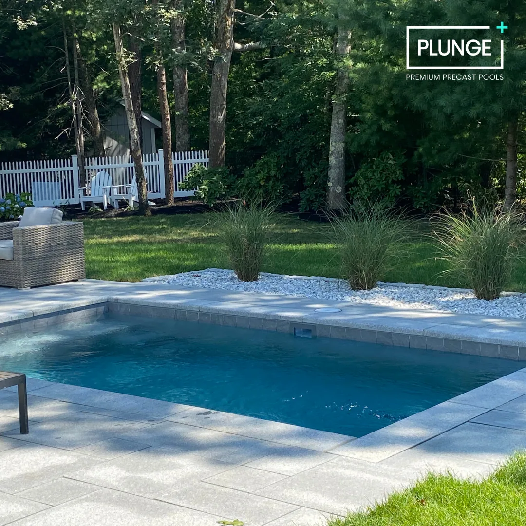 Backyard with a rectangular in-ground pool, surrounded by a stone patio, and landscaped with ornamental grass and trees. There are outdoor chairs and a white fence in the background.