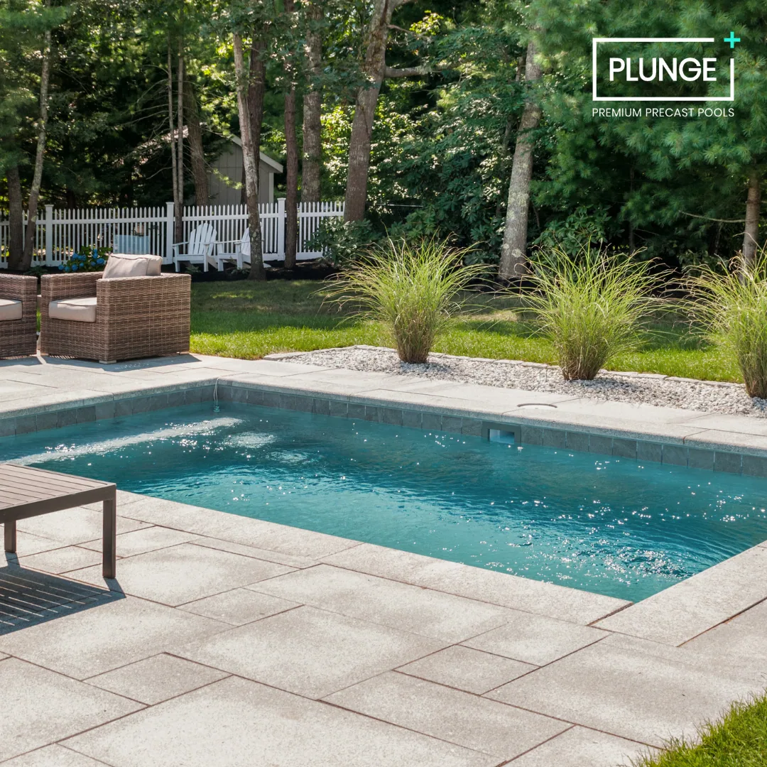 Backyard with a small swimming pool, patio furniture, green plants, trees, and a white picket fence in the background.