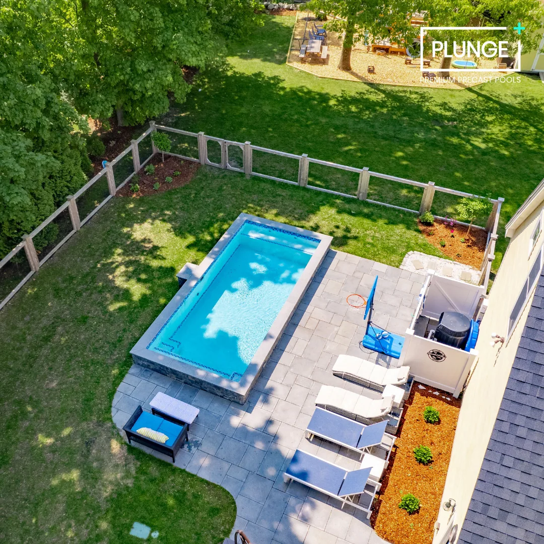 A backyard with a swimming pool, several lounge chairs, and a fenced grassy area with trees and a small garden.