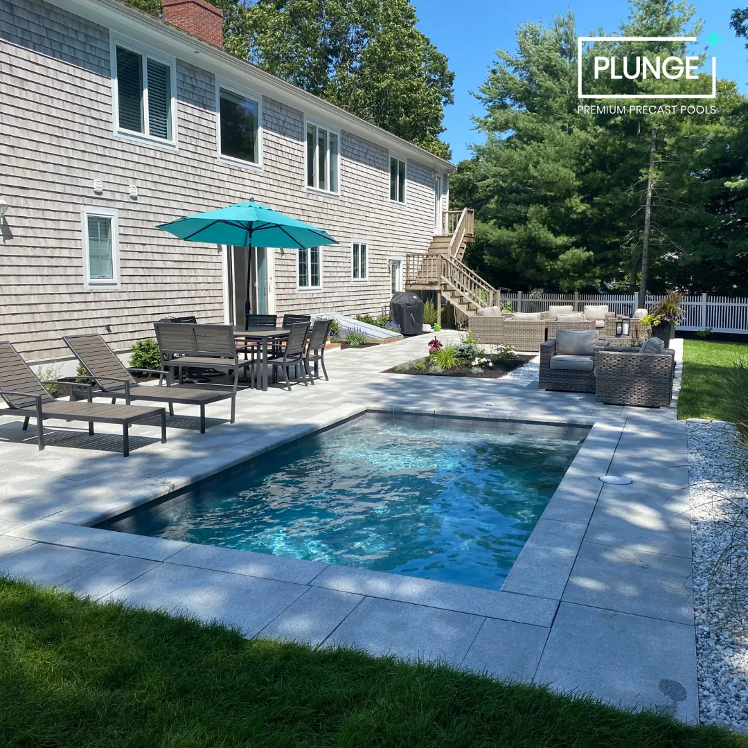 A backyard with a small rectangular pool, outdoor furniture including lounge chairs, a table with a blue umbrella, and a seating area with outdoor couches, surrounded by a white fence and greenery.