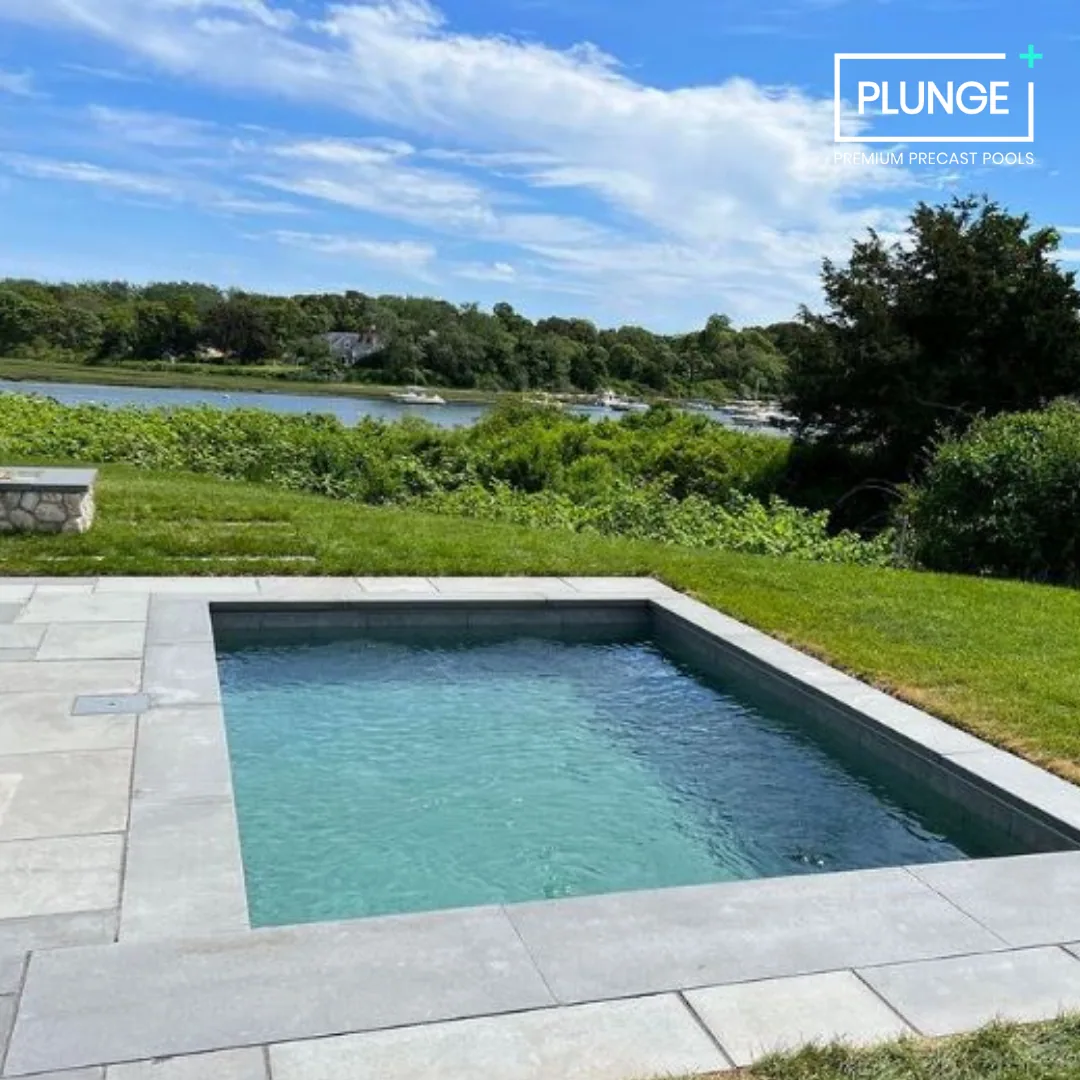 A modern rectangular pool with light gray stone decking, overlooking a lush green landscape with water and trees under a blue sky with some clouds.