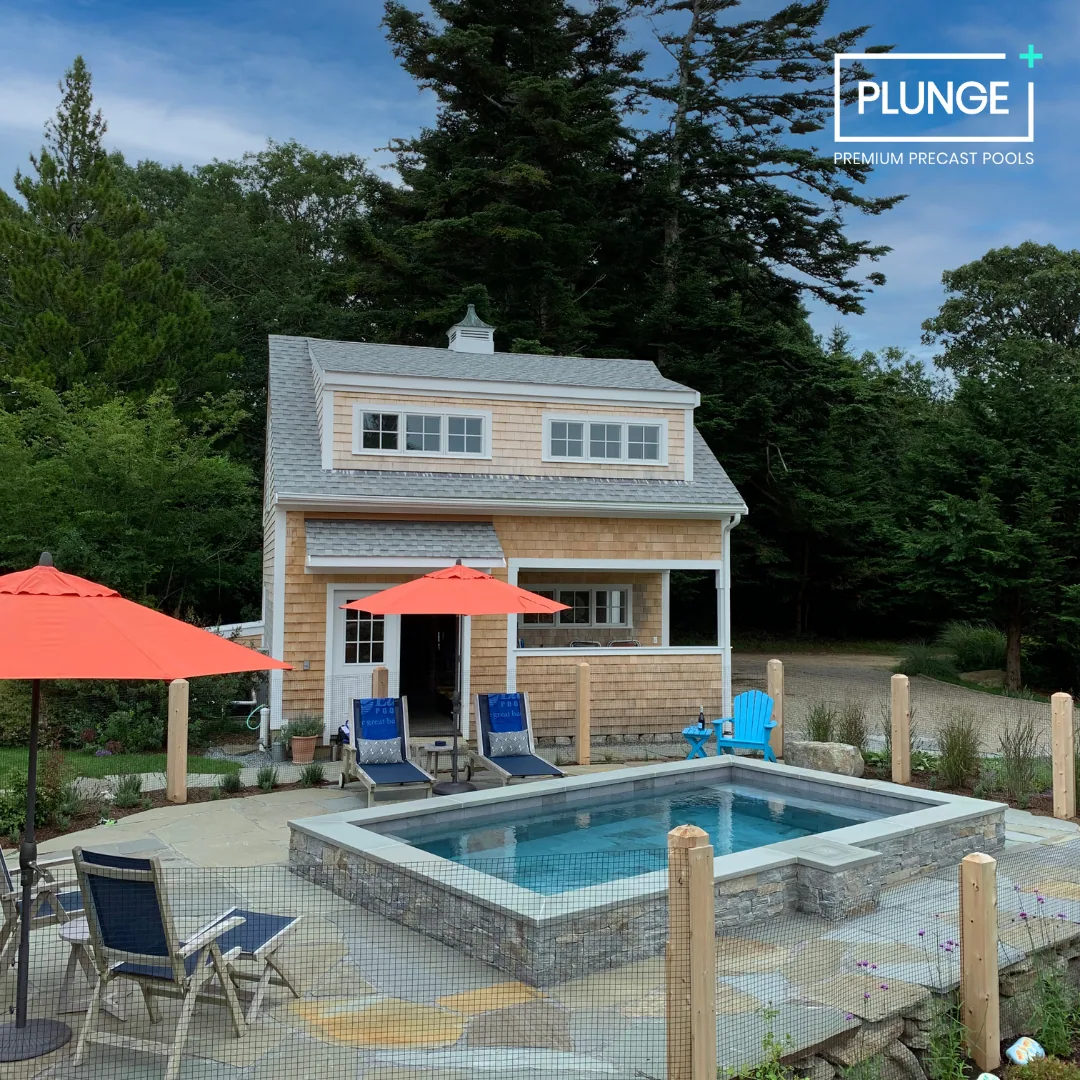 A backyard with a small pool surrounded by a stone deck, two lounge chairs with blue cushions and red umbrellas, a blue chair near the pool, and a two-story house with a porch, set amid trees against a partly cloudy sky.