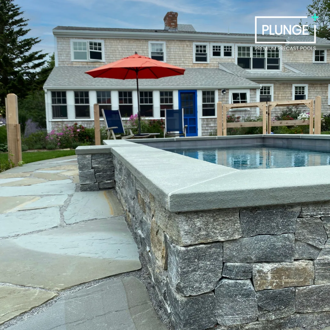 Backyard with a stone swimming pool, patio, and a house in the background featuring multiple windows, a blue door, and an umbrella with outdoor chairs.