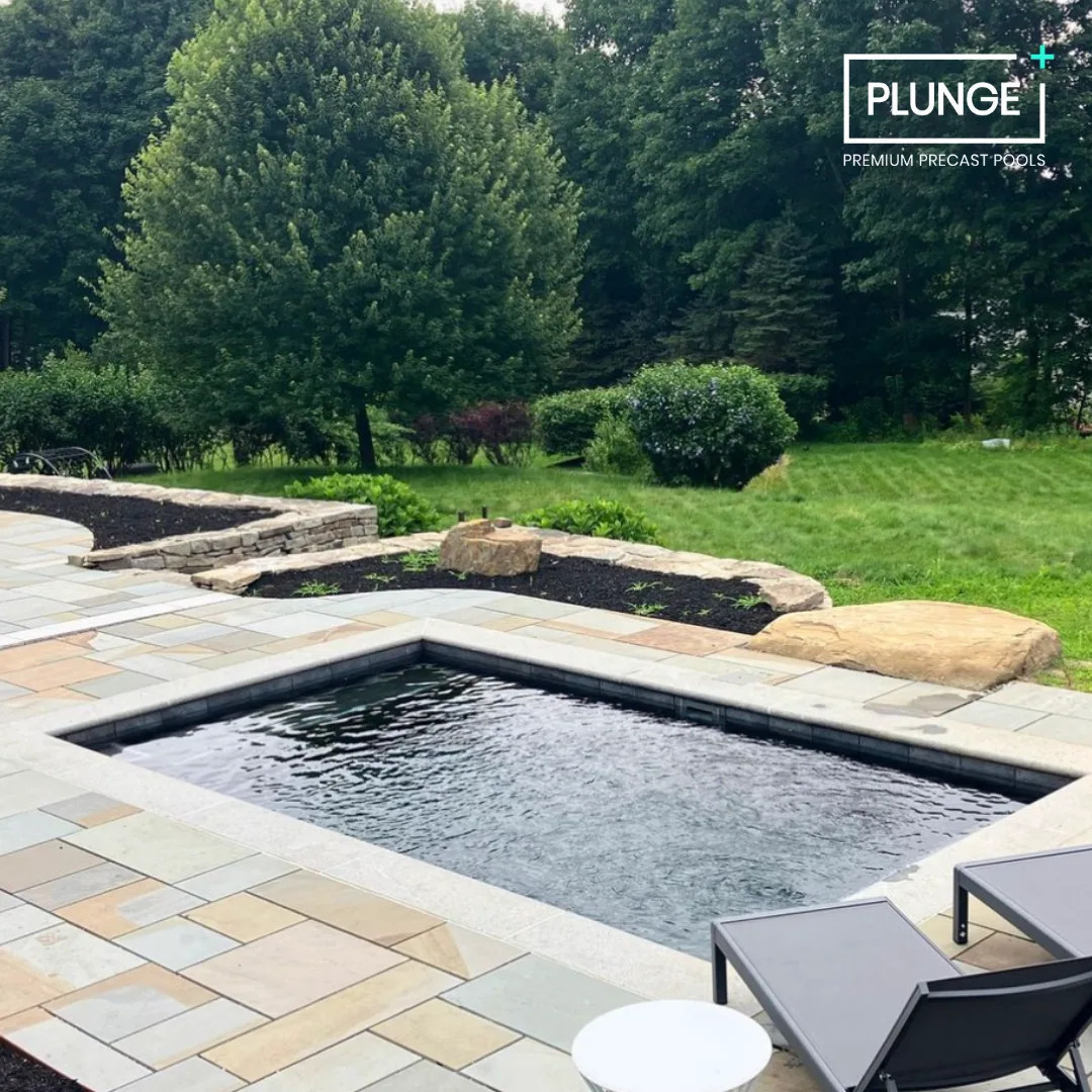 Backyard with a small rectangular pool, stone patio, green lawn, bushes, and trees.