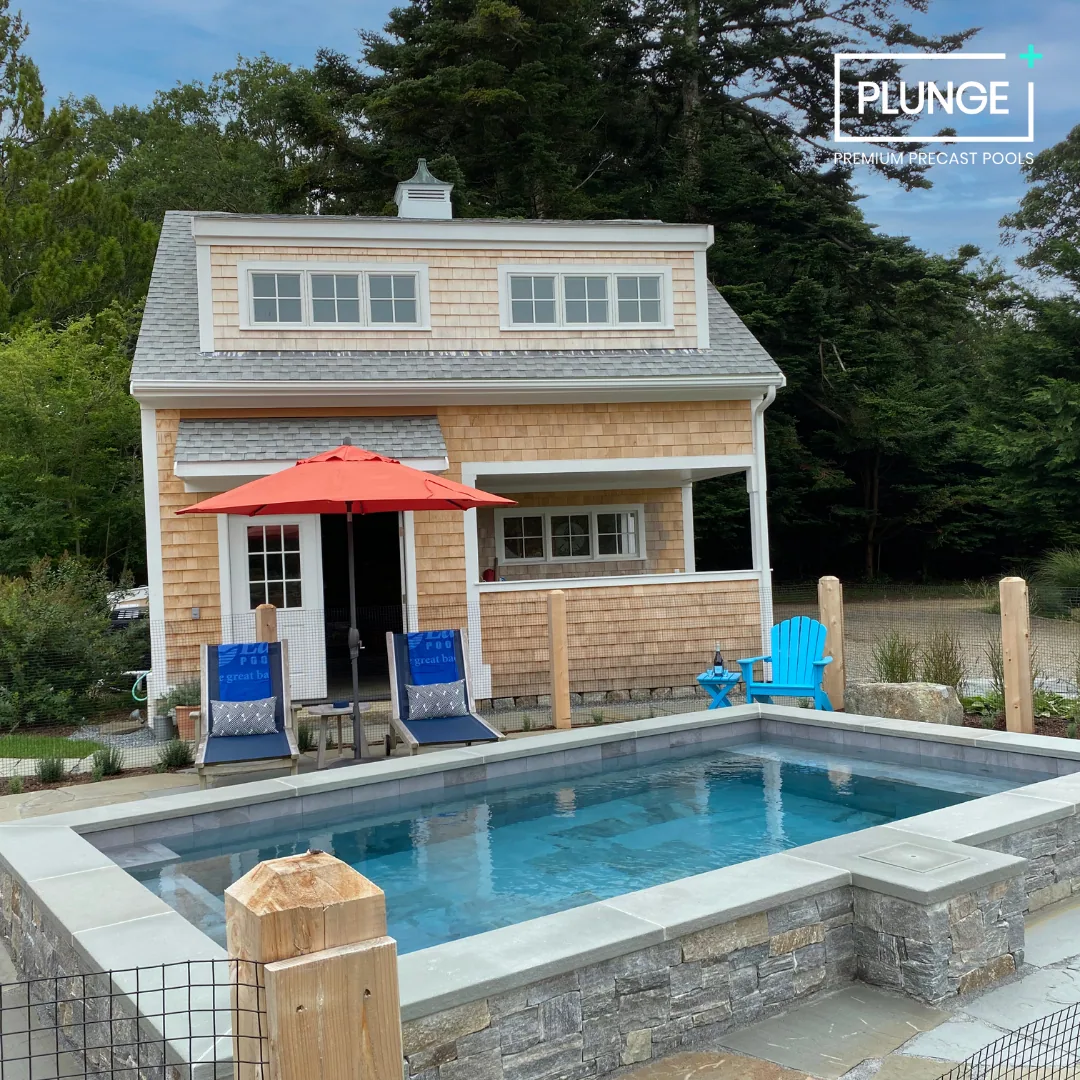 A backyard with a small rectangular pool, a wooden house with shingle siding, a red patio umbrella, blue lounge chairs with cushions, a blue Adirondack chair, and lush green trees in the background.