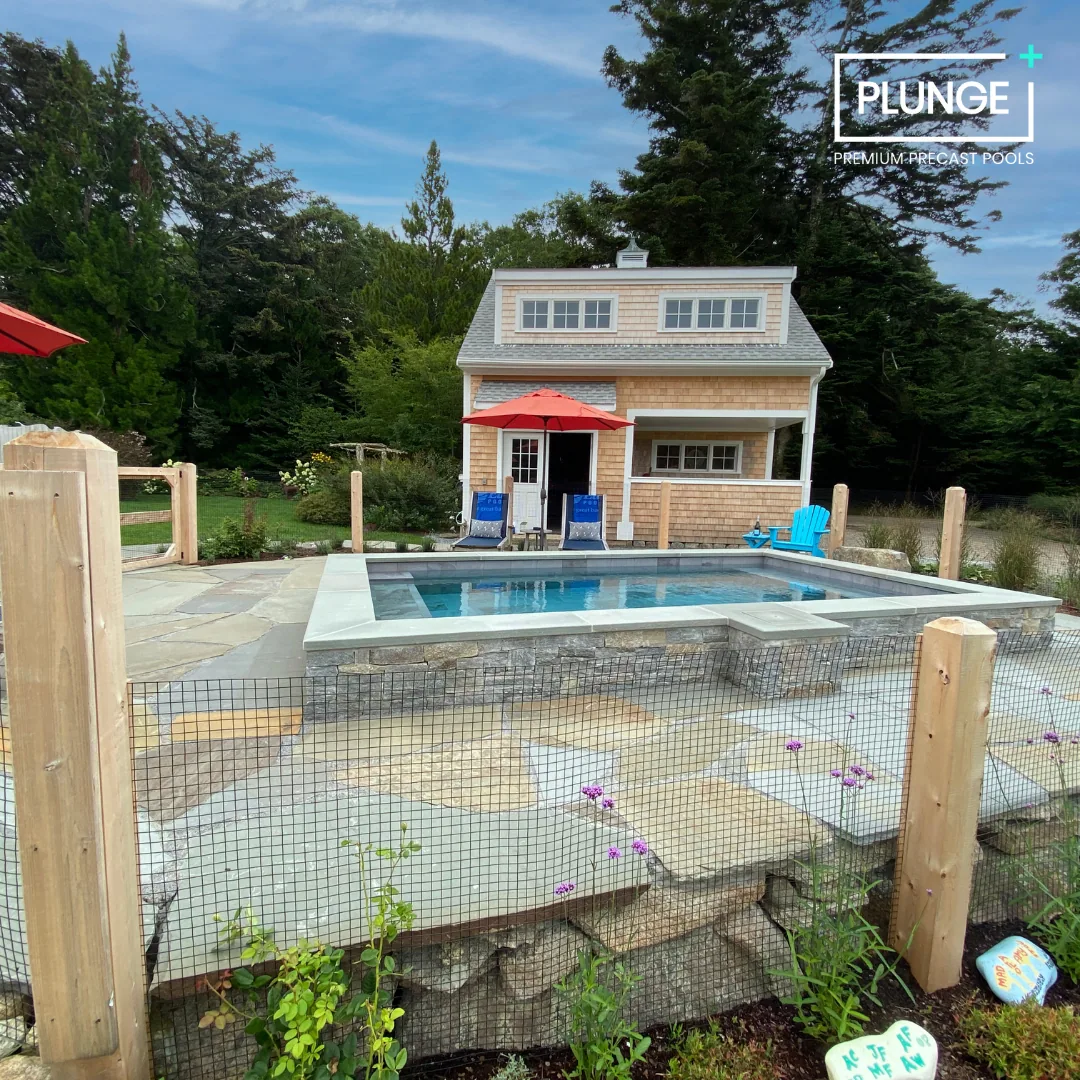 A backyard with a small in-ground pool surrounded by a stone patio, a two-story house with shingle siding, and outdoor furniture including blue chairs and a red umbrella. There are trees and green foliage in the background.