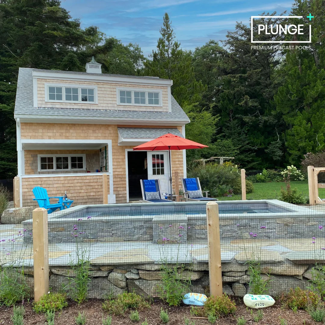 A backyard scene with a small house and a pool, surrounded by greenery and trees. The pool is enclosed by a wooden fence with rocks at the base, and there are blue chairs and an orange umbrella in the yard.