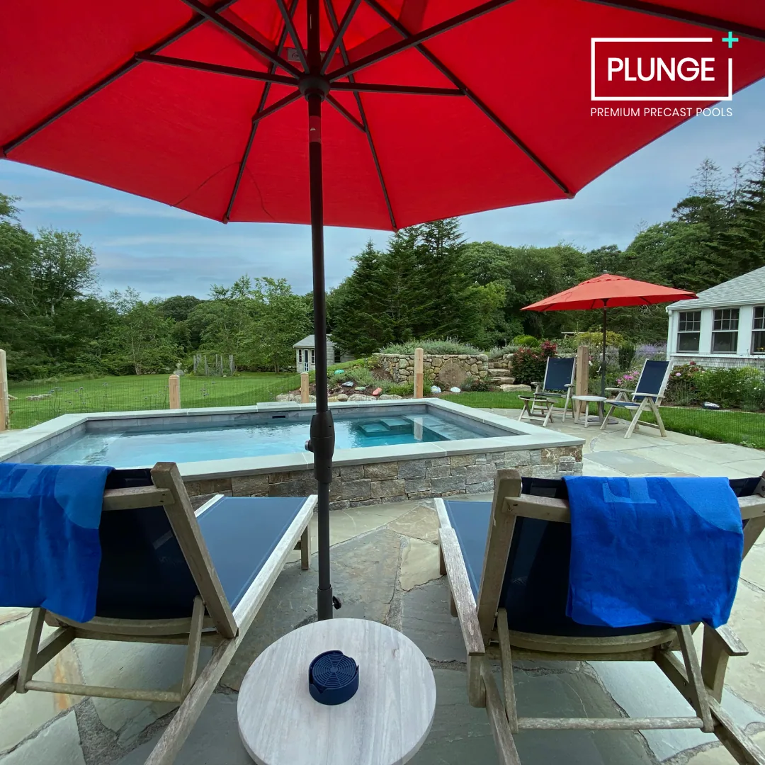 View of a backyard scene with a small pool, two lounge chairs with blue towels, a red patio umbrella, a small round table, and greenery in the background.