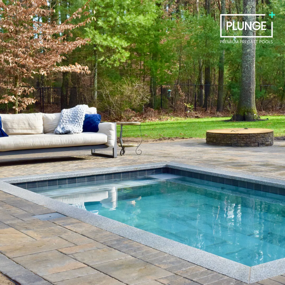 An outdoor backyard scene featuring a small rectangular pool with a stone border, a beige sofa with white and blue cushions, a small metal side table, a fire pit surrounded by a stone wall, and a wooded area with tall trees in the background.