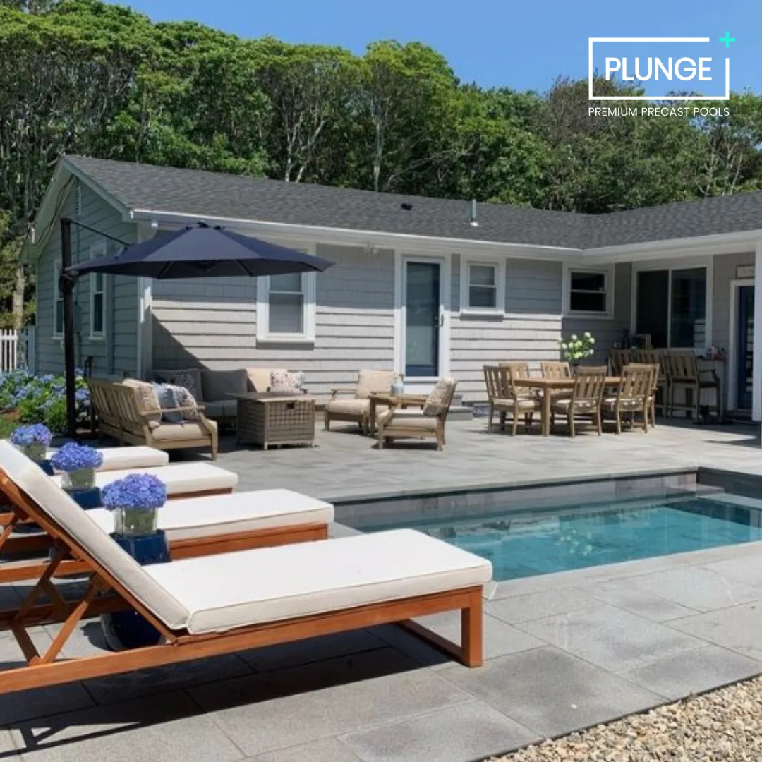 Outdoor backyard with a small pool, lounge chairs with white cushions, a patio with wooden furniture, a patio umbrella, and a house with gray siding and multiple windows, surrounded by green trees.