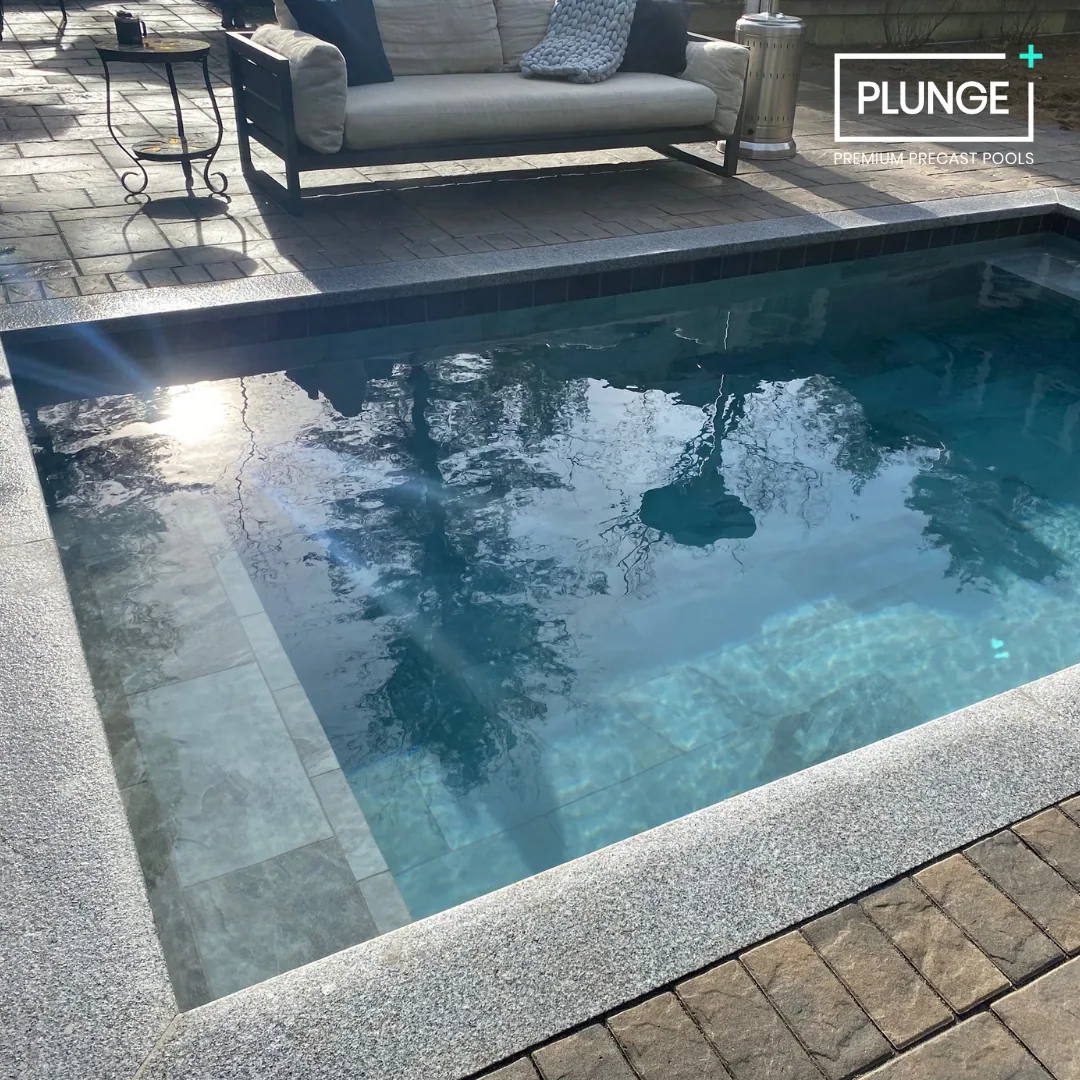 An outdoor backyard with a small rectangular plunge pool, a lounge sofa with pillows, a small table, and some patio furniture on a paved surface.