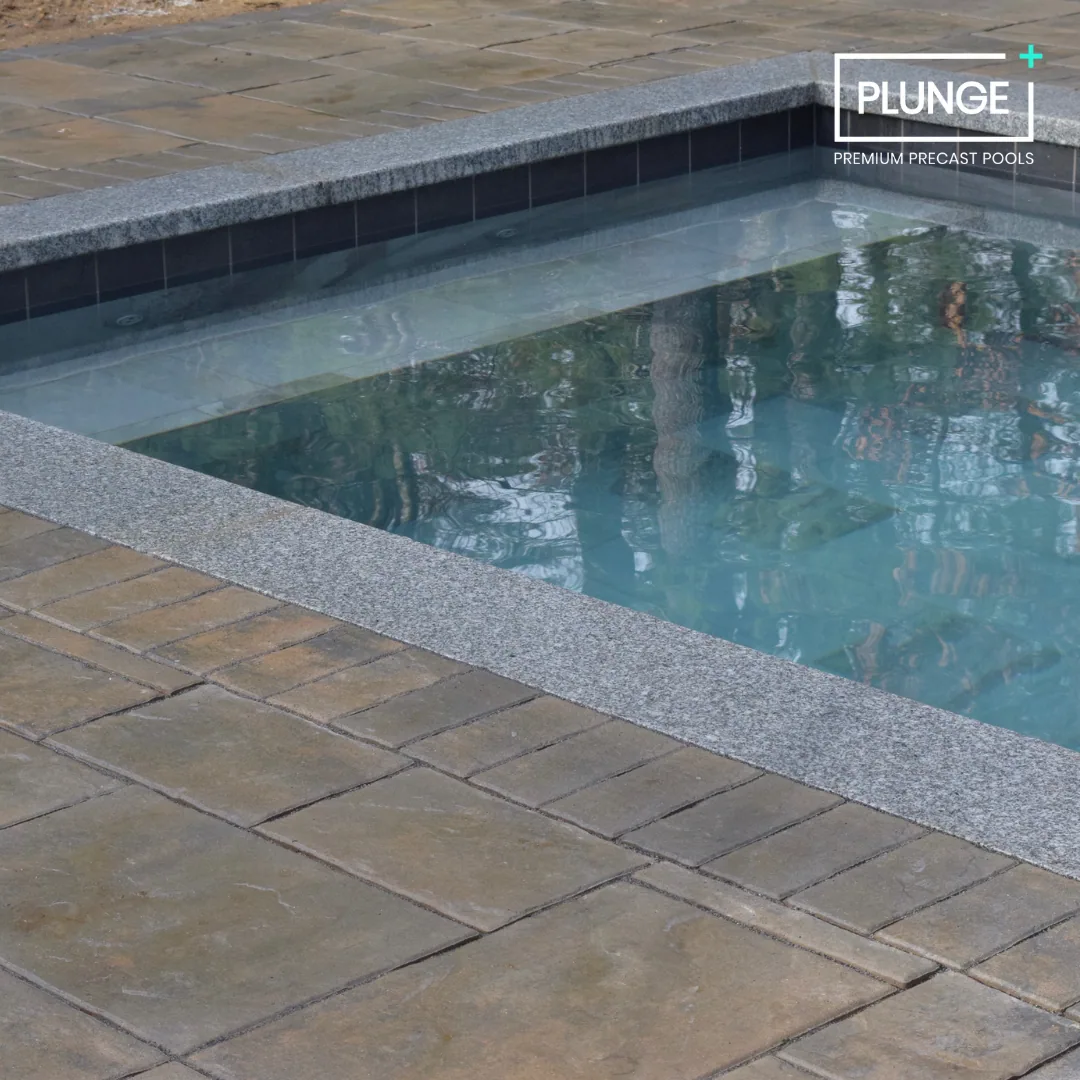 Close-up of a rectangular concrete and stone pool with clear water, surrounded by beige stone tiles and a gray stone border, with a logo that says "PLUNGE PREMIUM PRECAST POOLS" in the upper right corner.