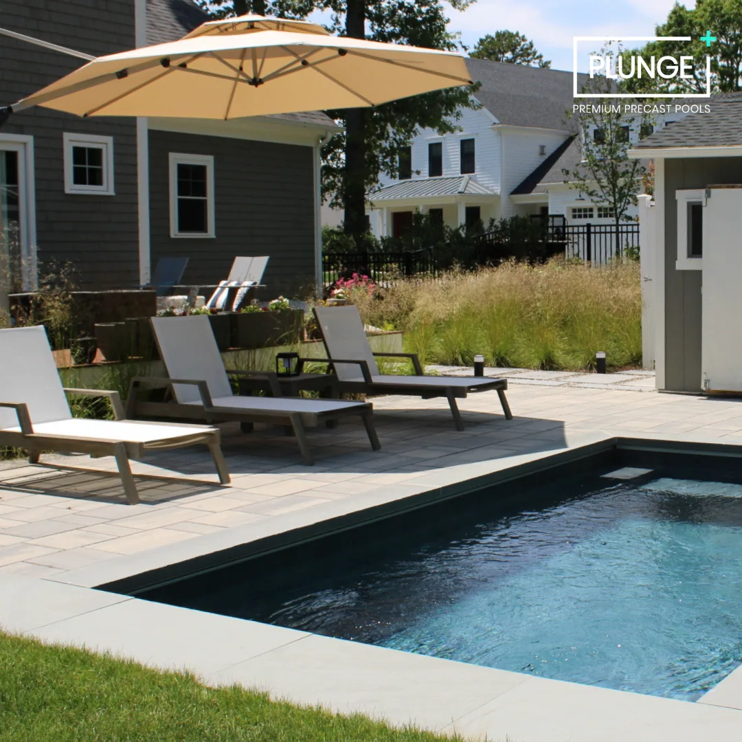 A backyard with a small in-ground swimming pool, lounge chairs, and a large beige patio umbrella. There are plants and grass surrounding the area and neighboring houses in the background. The image includes a logo for 'PLUNGE Premium Precast Pools'.