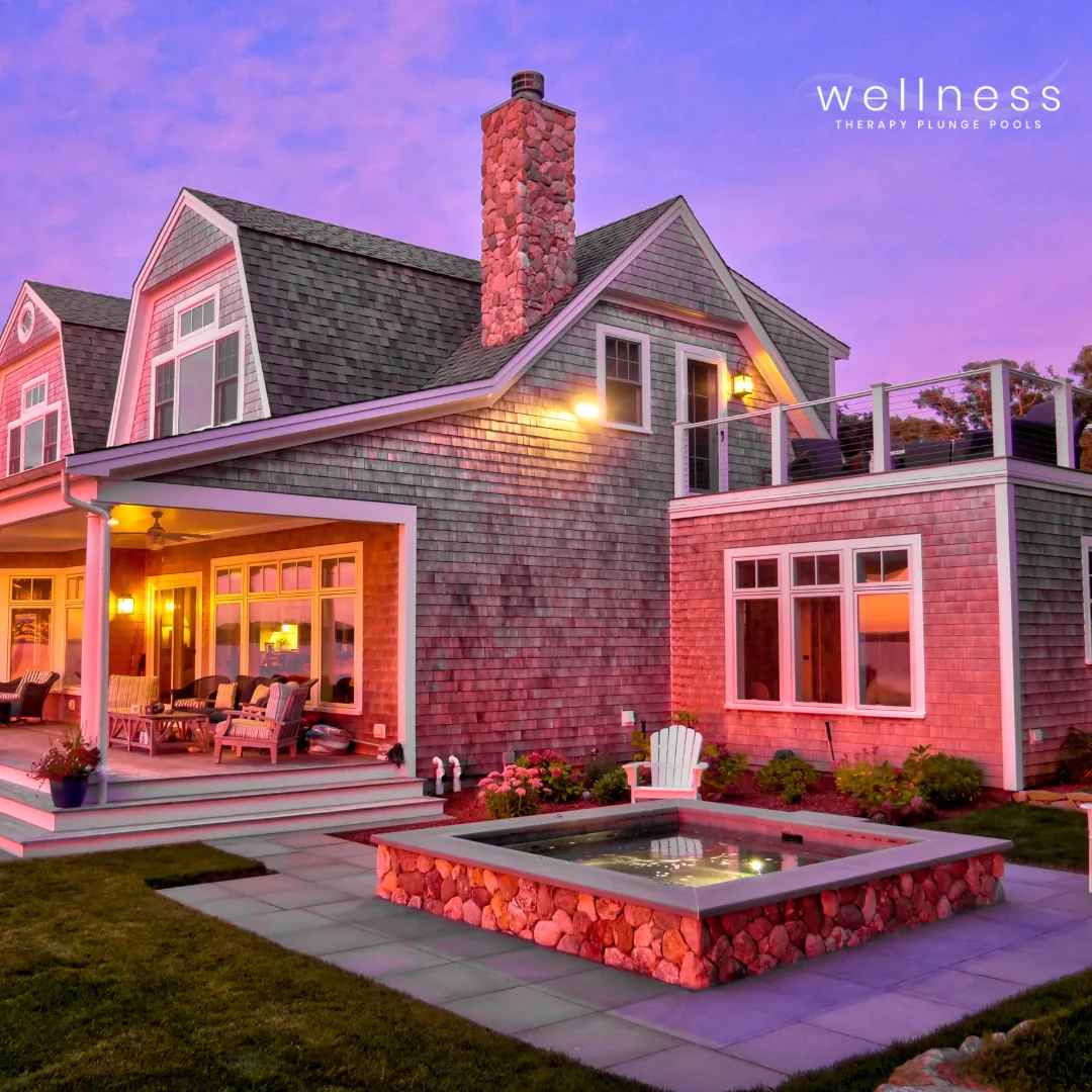 A house with gray shingled exterior, multiple gabled roofs, a stone chimney, a porch with outdoor furniture, a small pond with a stone border, and a backyard with flower beds. Sunset sky with pink and purple hues.