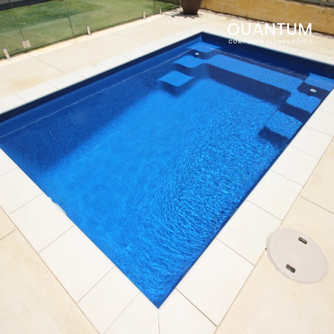 A modern blue rectangular pool with integrated steps, surrounded by a white tiled deck and glass fencing, with a potted plant in the background.