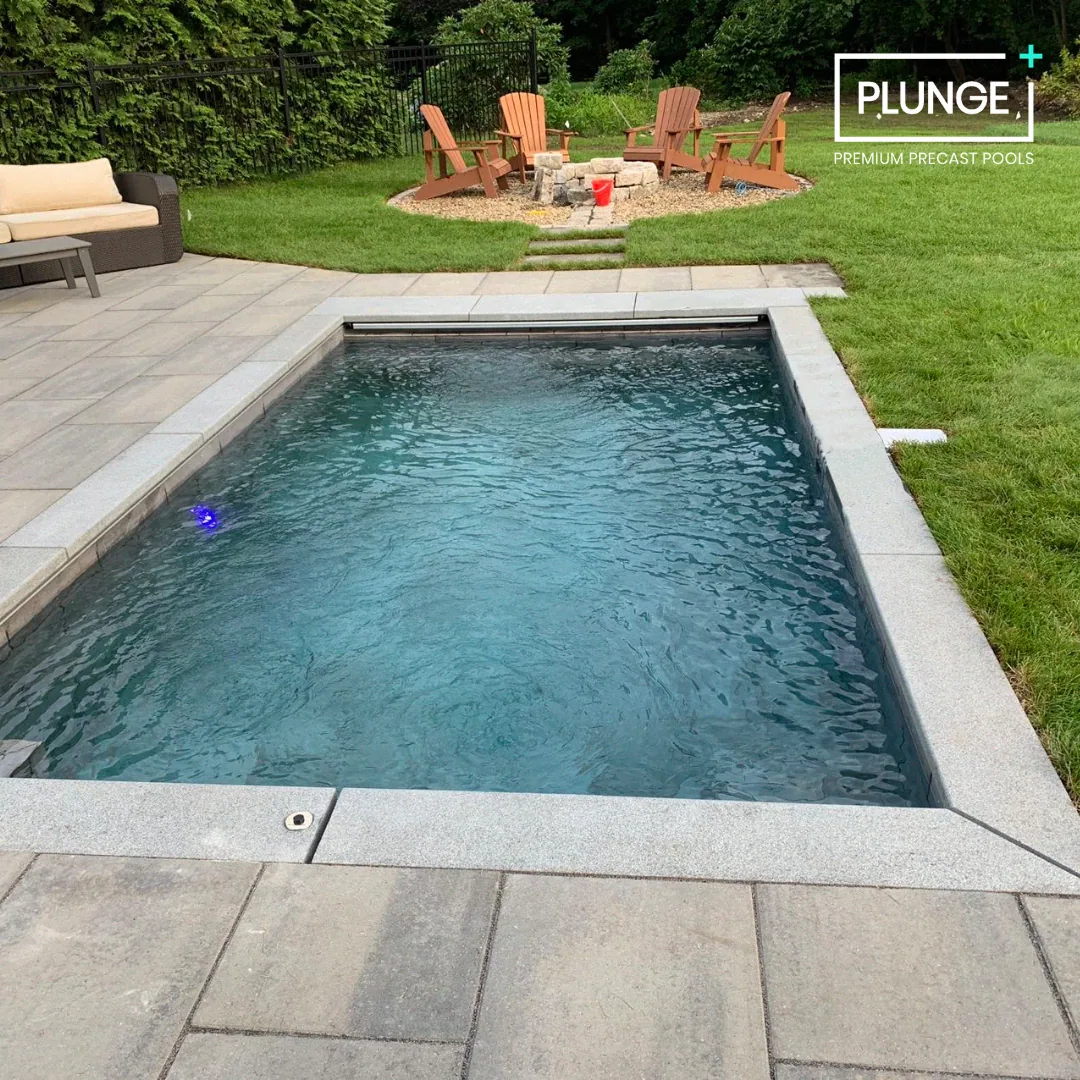 A backyard with a small rectangular pool with a gray stone border, a stone fire pit area with wooden chairs, a green lawn, and a paved patio with a couch and a table.