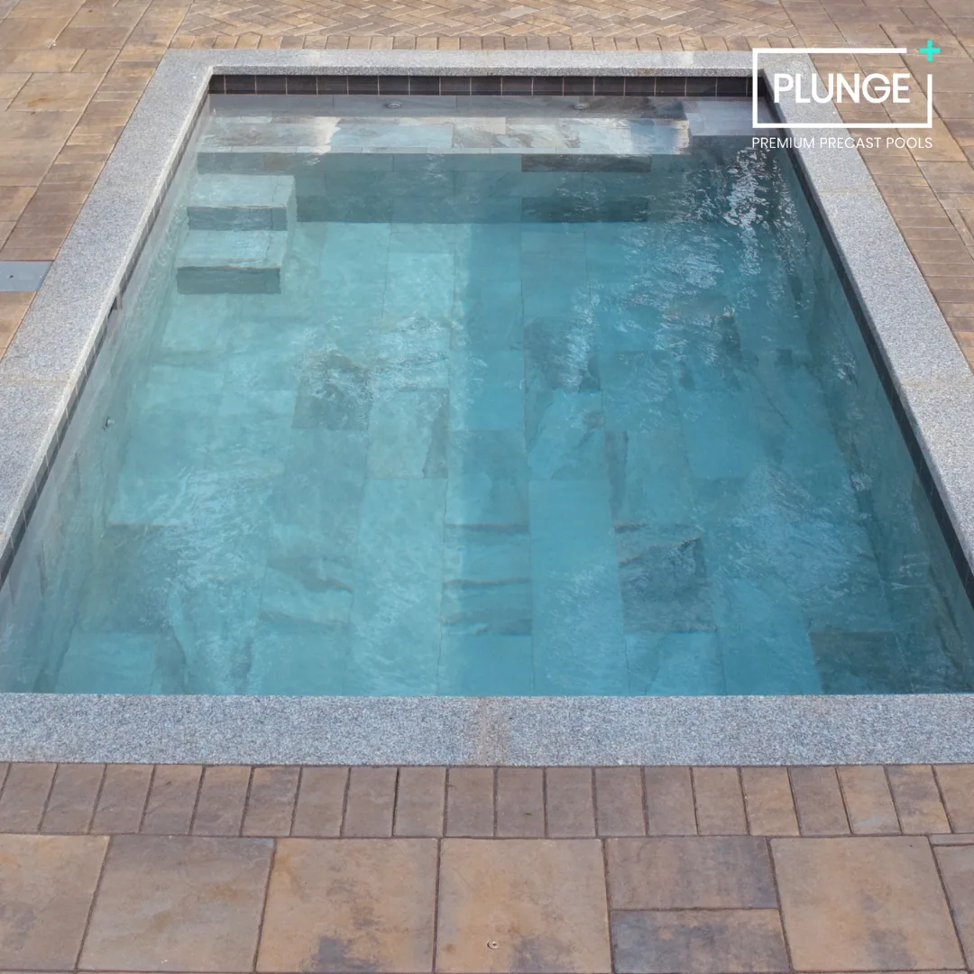 A rectangular in-ground precast swimming pool with steps in the shallow end, surrounded by a stone tile deck with a border of smaller tiles, and the pool water is clear and blue.