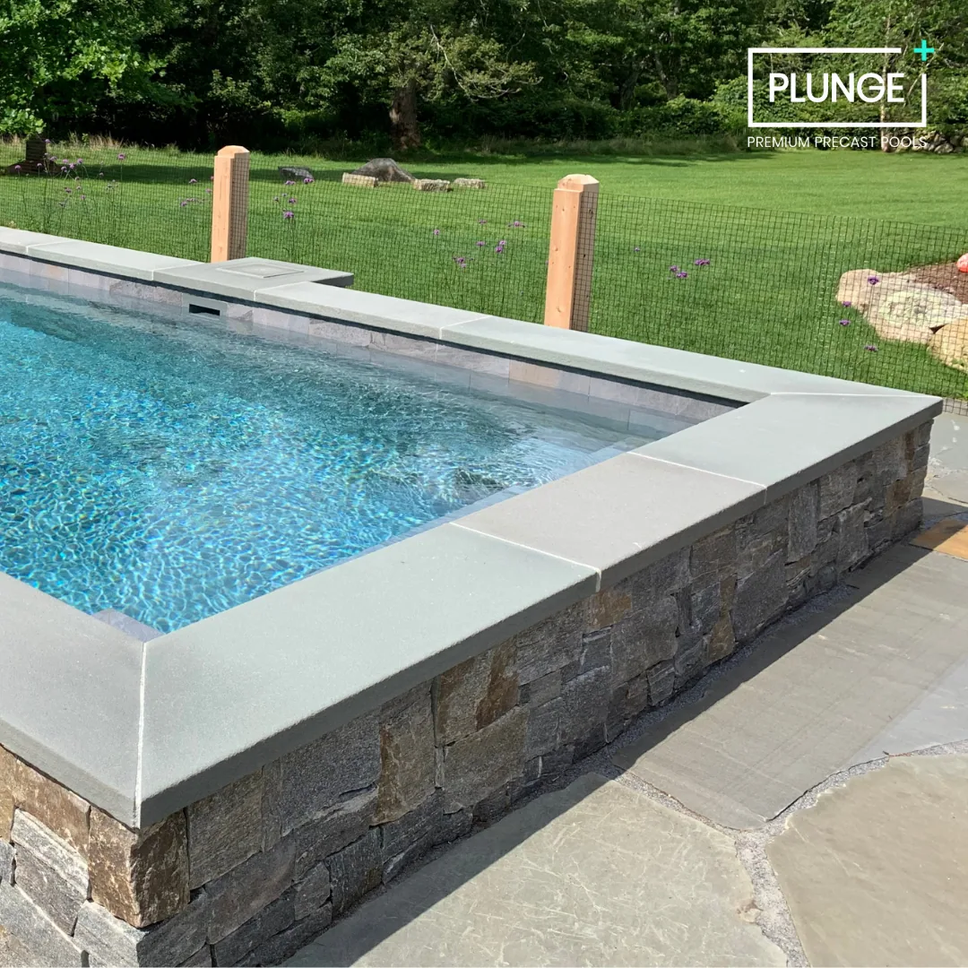 A rectangular in-ground swimming pool with blue water, a stone and concrete border, and a surrounding concrete patio, next to a grassy yard enclosed by a wire fence with wooden posts, and trees in the background.