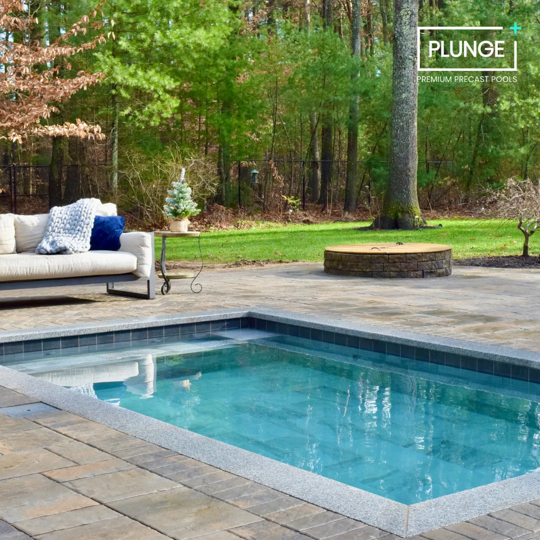 Backyard with a rectangular in-ground swimming pool, outdoor sofa with pillows, small Christmas tree on a side table, stone fire pit, and lush green trees in the background.