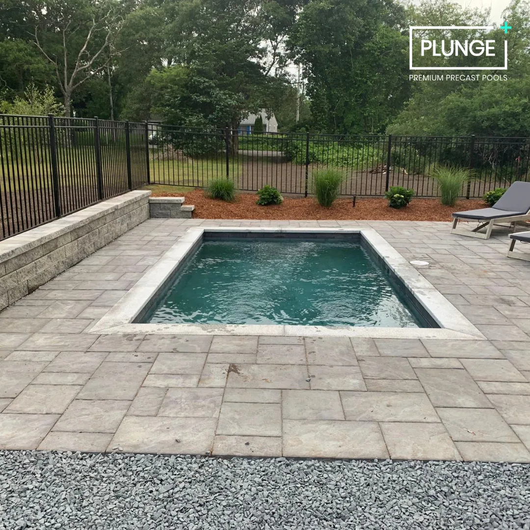 A small rectangular plunge pool with a stone border, surrounded by a paved patio area and black metal fencing. There are lounge chairs on the right side, plants and shrubs in the background, and trees beyond the fence. The image has a logo in the top