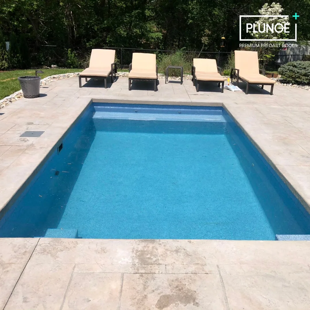 A rectangular swimming pool with blue water, surrounded by a stone patio. Four lounge chairs are positioned along the back edge, with a small table between the second and third chairs. A trash can is on the left side of the patio. There is a fence an