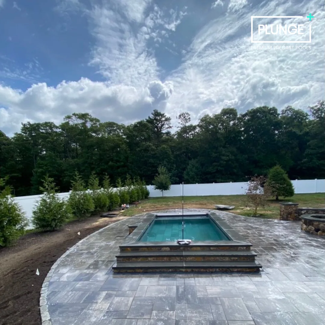 A backyard with a rectangular swimming pool on a stone patio, surrounded by trees and a white fence, under a partly cloudy sky.