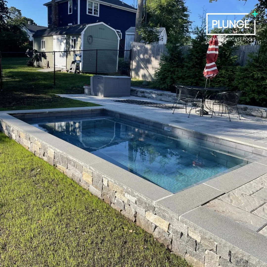 A backyard pool with clear water, surrounded by stone and brick paving, with a patio table and umbrella, green grass, a small shed, a fence, and trees in the background.