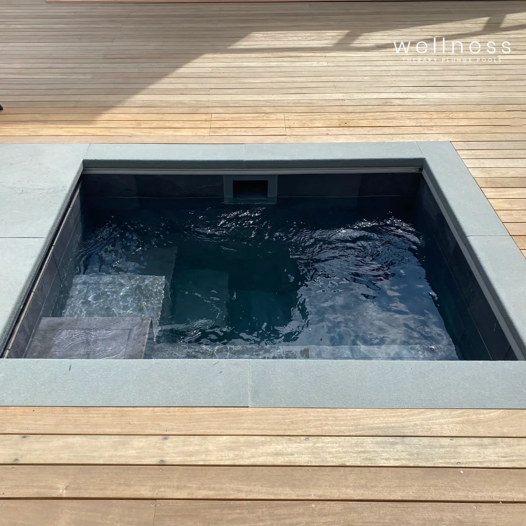 A small outdoor hot tub or spa with dark water, surrounded by light gray stone tiles and a wooden deck area.