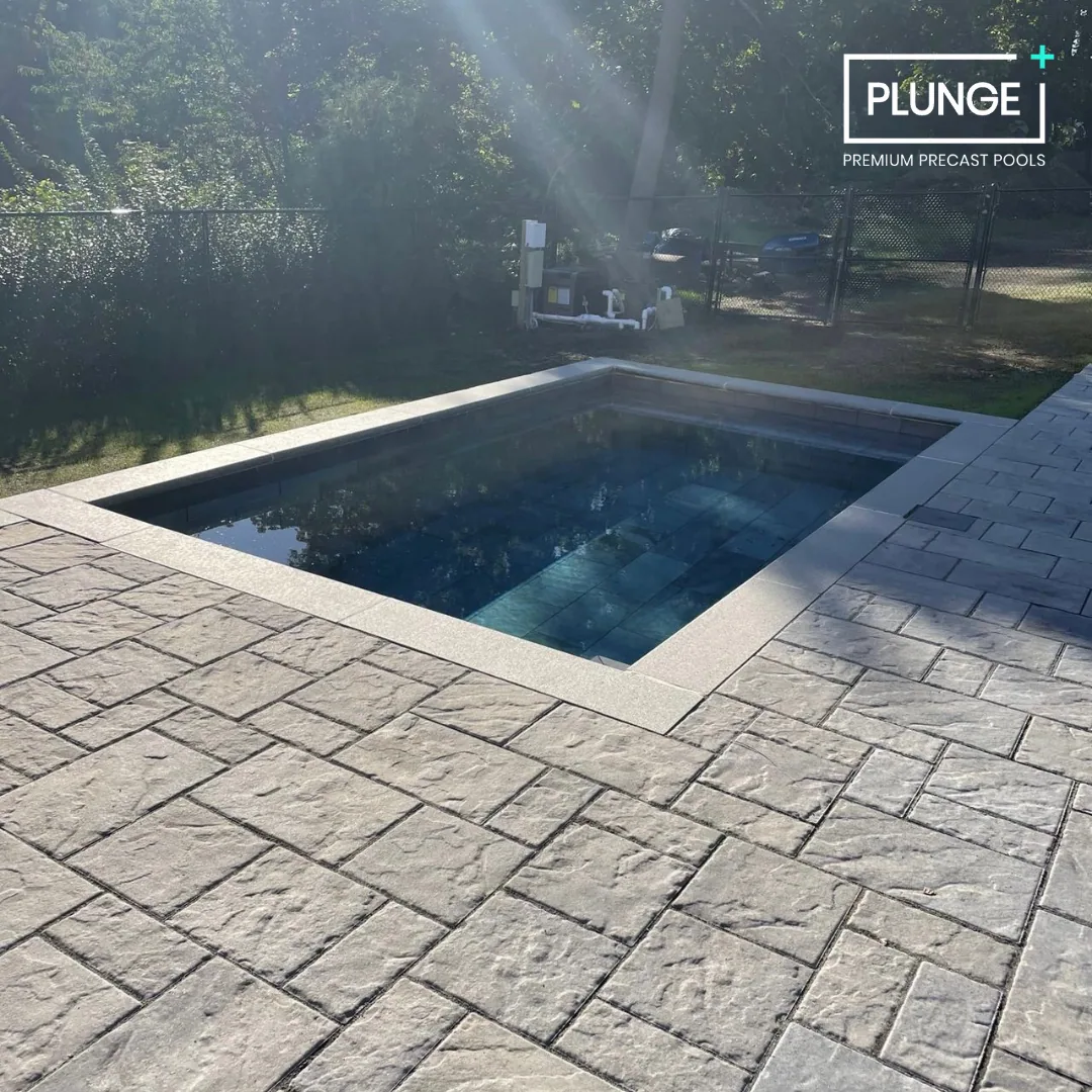 A rectangular inground pool with a stone border and concrete paving surrounding it, located outdoors with trees and a fence in the background.