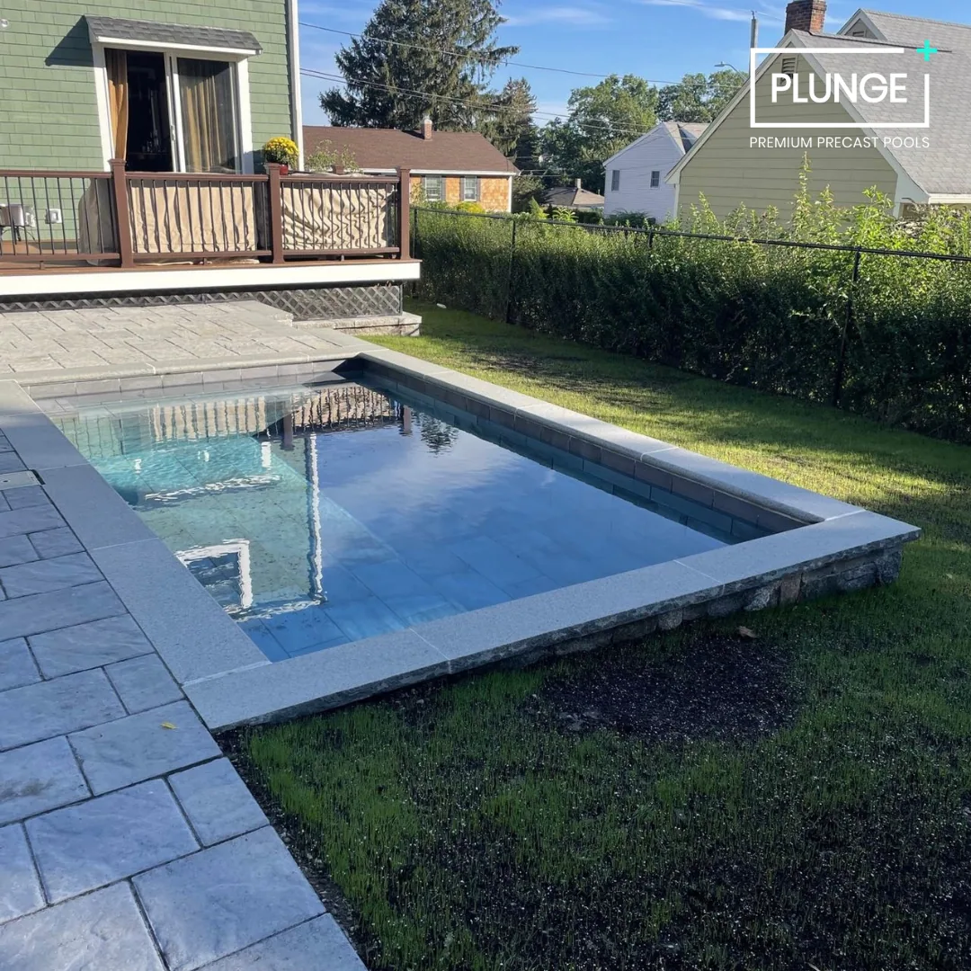 A backyard with a small rectangular in-ground pool, a stone patio, a deck with outdoor furniture, and a green house with a sliding door. There are neighboring houses, a hedge, and trees in the background.