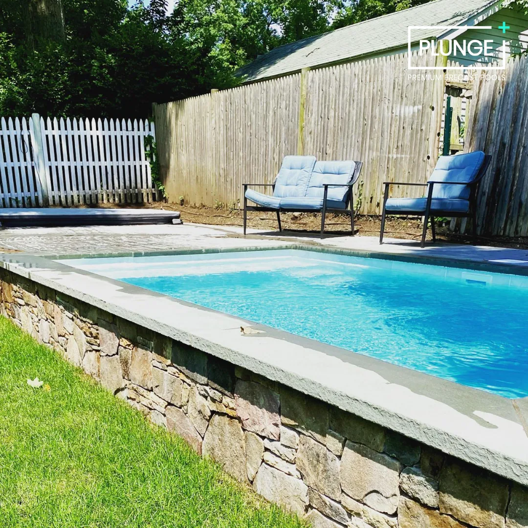 An outdoor swimming pool with a stone and concrete border, surrounded by a grassy yard, two cushioned chairs, and a wooden fence with trees in the background.