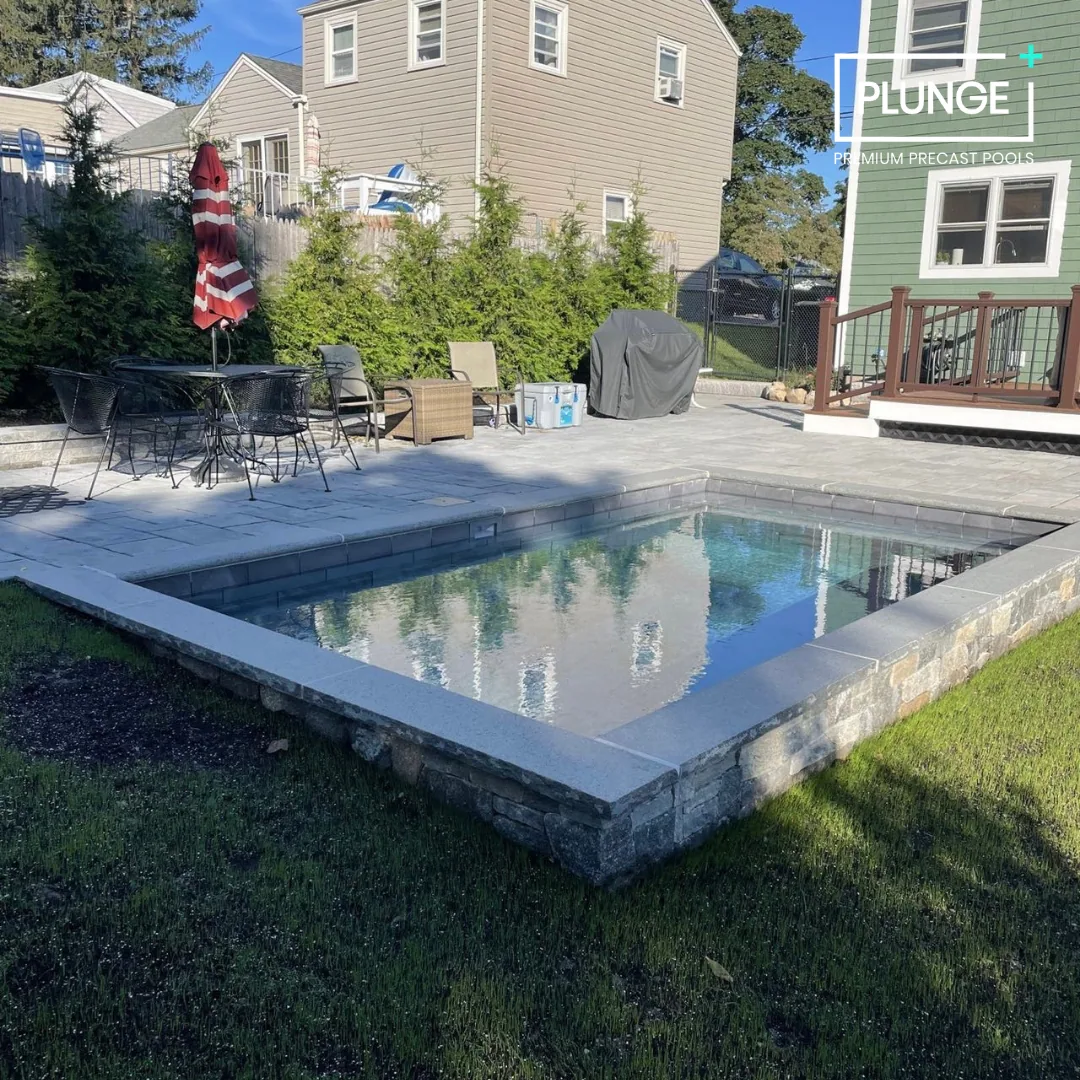 Residential backyard with a small pool, outdoor seating including a table with a red and white umbrella, a barbecue grill covered with a black cover, and two chairs. There are some coolers and chairs on the patio, with a house and trees in the backgr