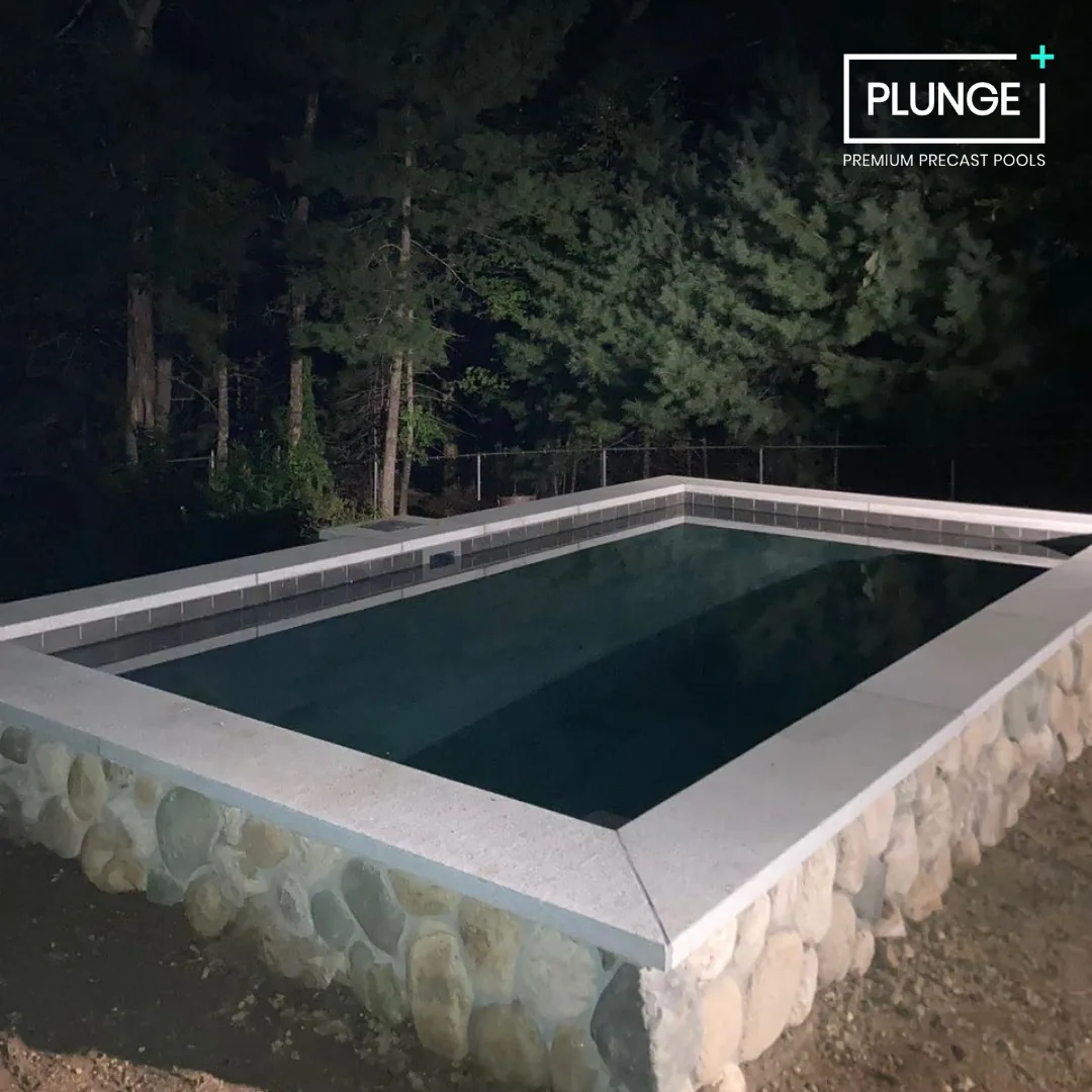 Night view of a rectangular, above-ground pool with stone and tile sides, surrounded by trees in the background.