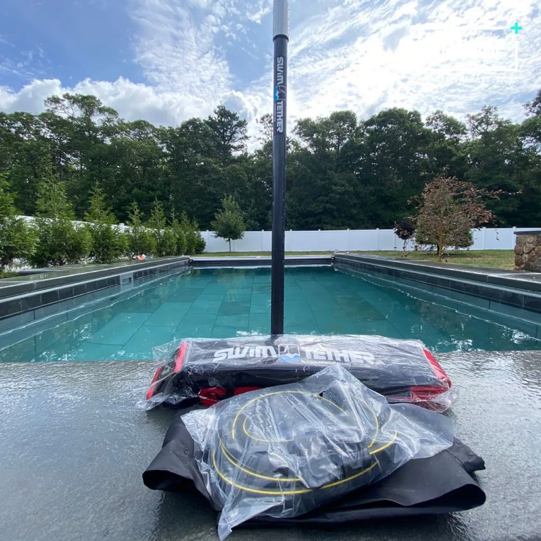 A swimming pool outdoors with a life jacket and other gear placed on the poolside in front of it, under a partly cloudy sky surrounded by trees and a white fence.