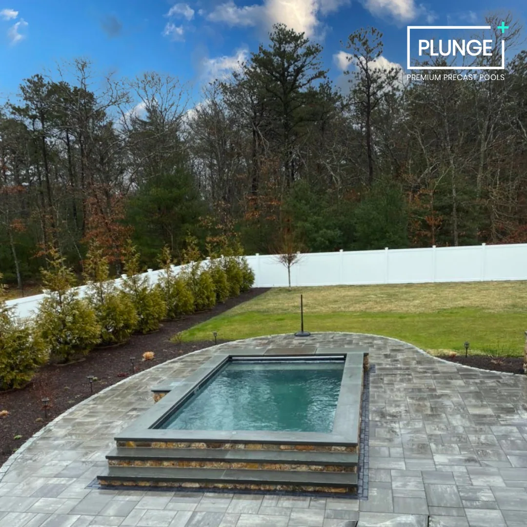 Backyard with a rectangular swimming pool on a paved patio, surrounded by a white fence, trees in the background, and a grassy yard.