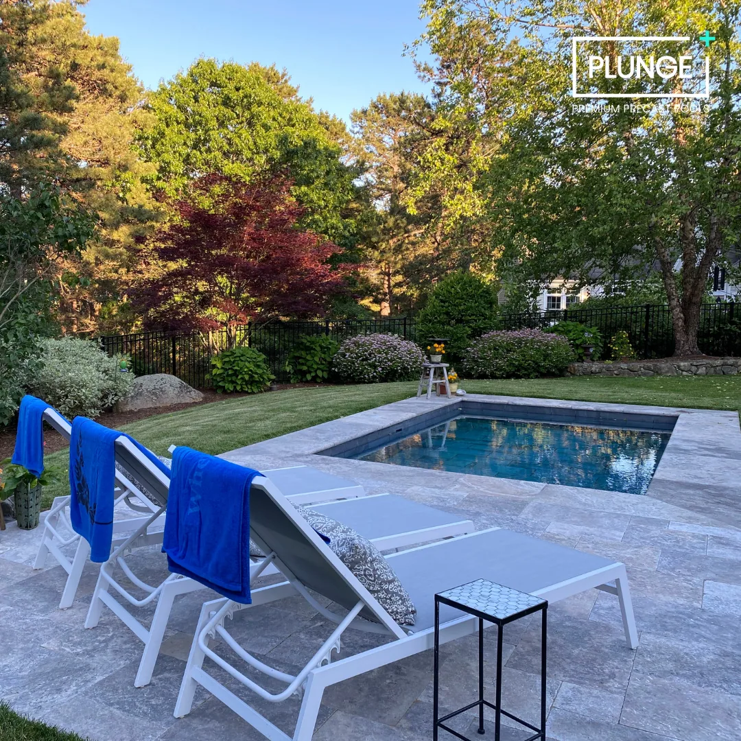 Backyard with a small swimming pool, four lounge chairs with blue towels, a small side table, and lush green trees and bushes in the background.