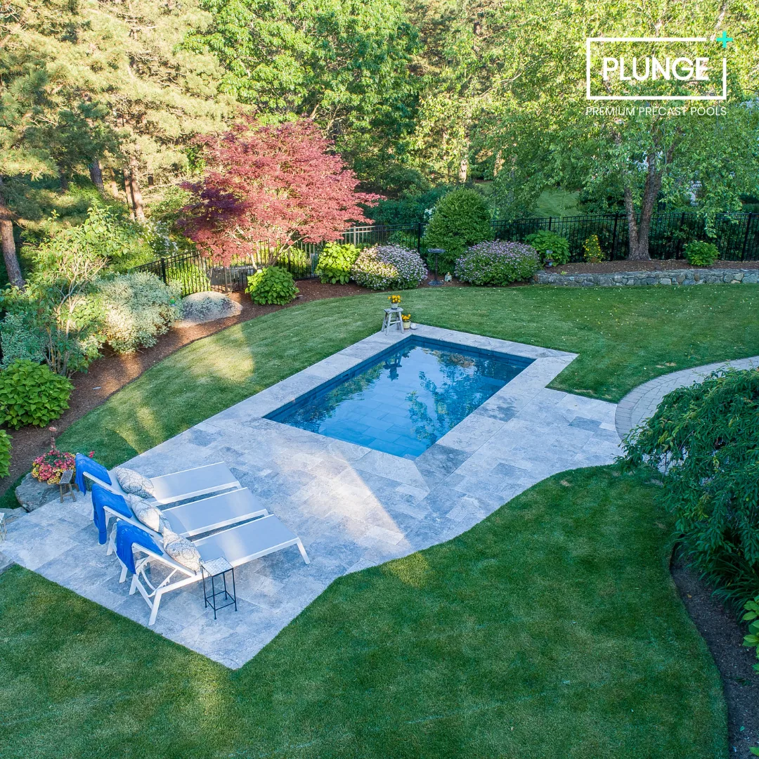 A backyard with a rectangular swimming pool surrounded by a stone patio, green grass, and landscaped bushes and trees.