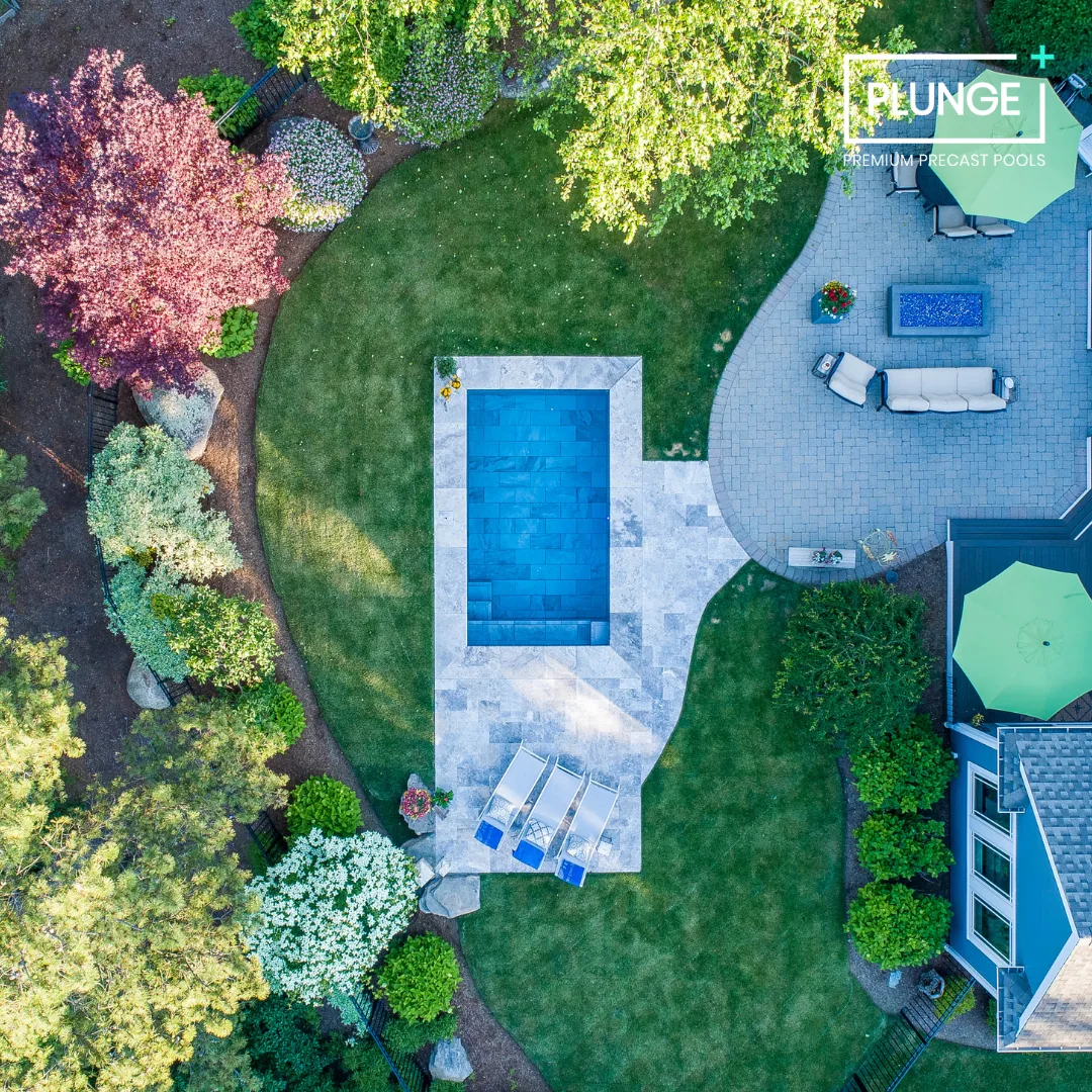 A backyard with a rectangular pool surrounded by a stone patio, green lawn, trees, and garden beds. There are lounge chairs and outdoor furniture with umbrellas, along with a small fire pit and decorative plants.