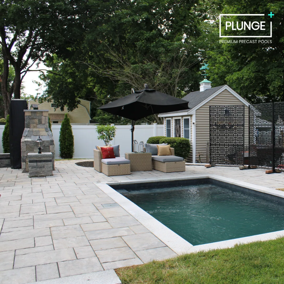 Backyard with a small pool, outdoor seating with cushions and a large black umbrella, stone patio, white fence, small shed, and trees in the background.