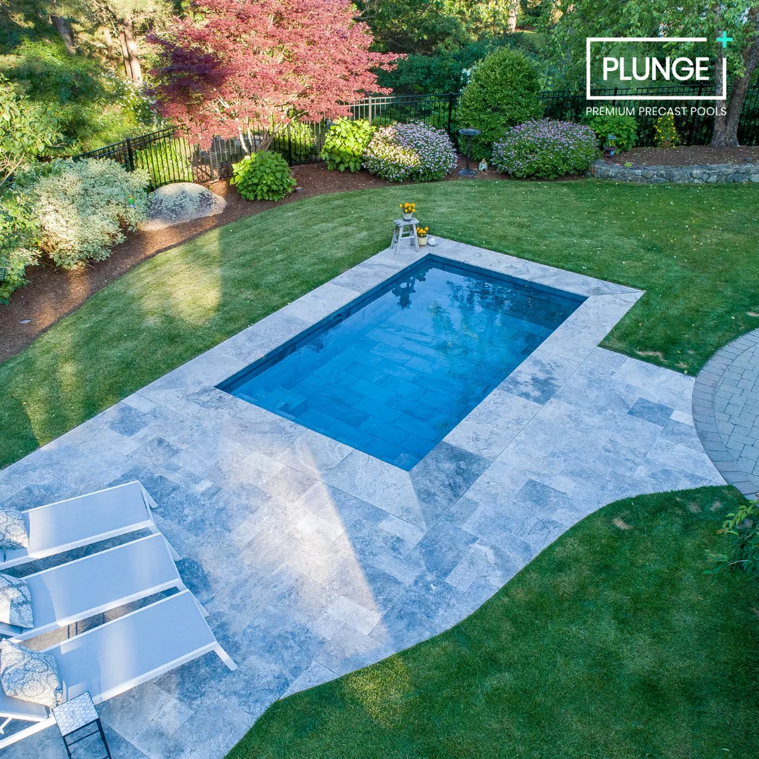 Backyard with a rectangular blue swimming pool surrounded by gray stone patio, green grass, trees, and flowering bushes, with lounge chairs and a small table.
