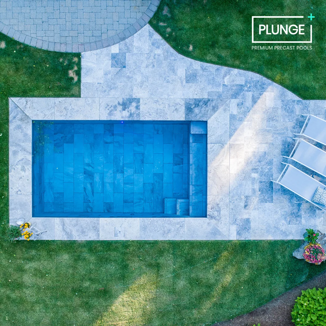 An overhead view of a rectangular blue swimming pool surrounded by stone paving and green grass, with white lounge chairs on the right side and small flower bushes in the corners.