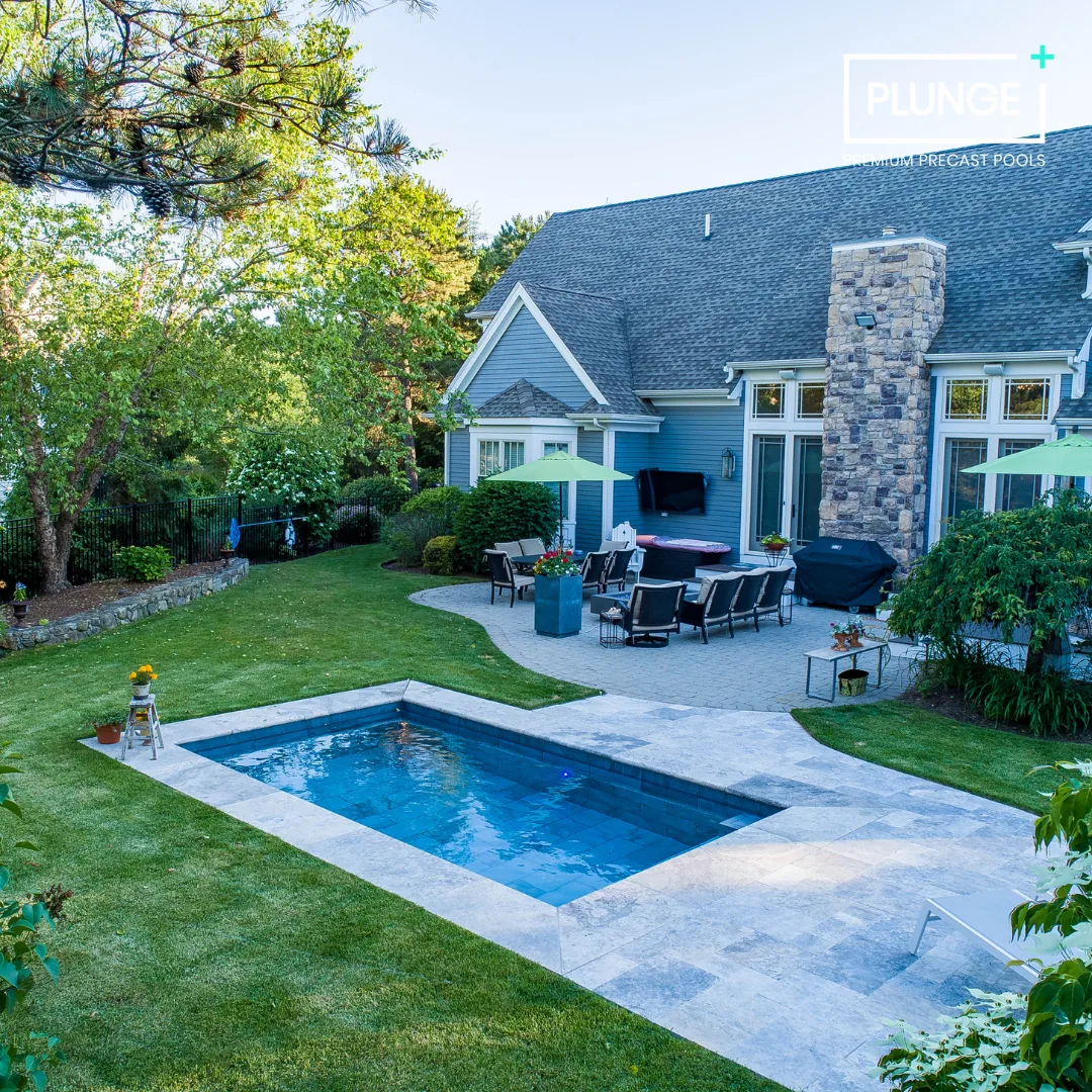 Backyard with a swimming pool, patio furniture with umbrellas, and a house with a stone chimney, surrounded by trees and greenery.