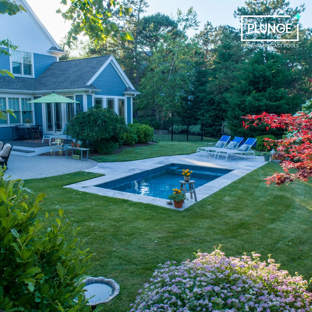 Backyard with in-ground swimming pool, poolside lounge chairs, patio furniture, lush green grass and trees, and a blue house with a patio umbrella.