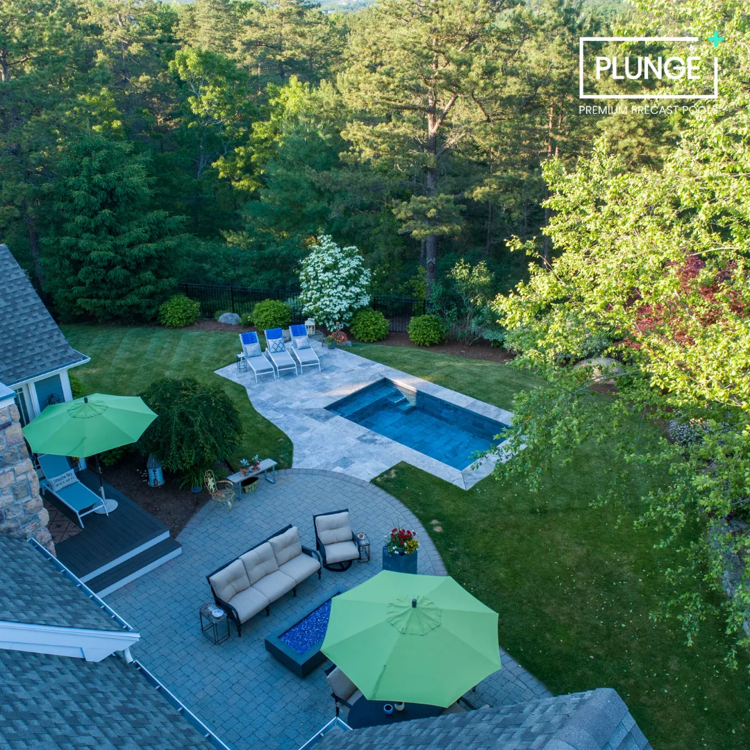 A backyard scene with a swimming pool, green lawn, outdoor seating area with beige couches and chairs, green umbrellas, a small patio with a table, lawn chairs, and a garden with trees and shrubs.