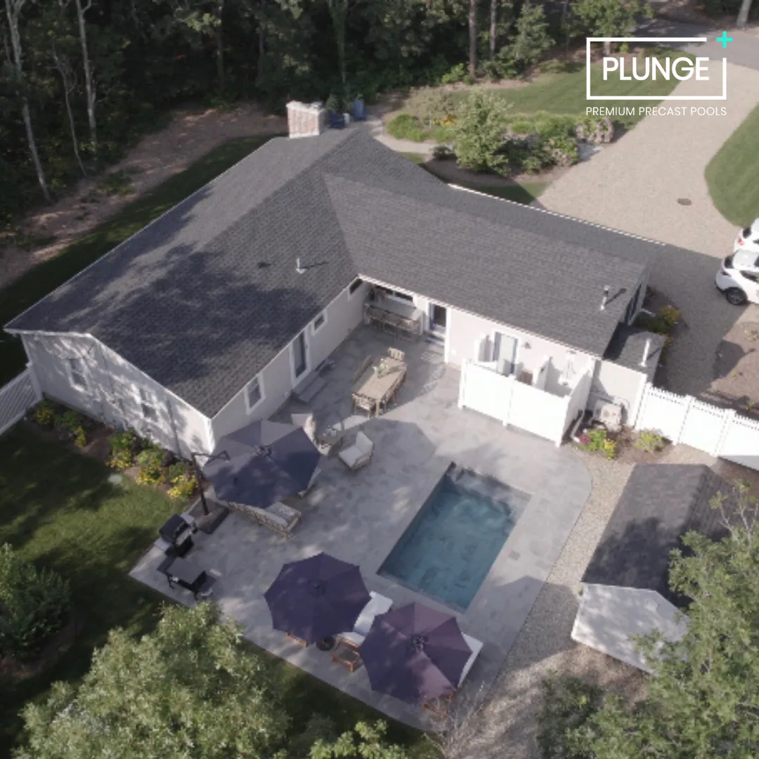 Aerial view of a backyard with a swimming pool, patio furniture, and umbrellas, attached to a house with a gray roof. There are trees and a driveway with parked cars nearby.