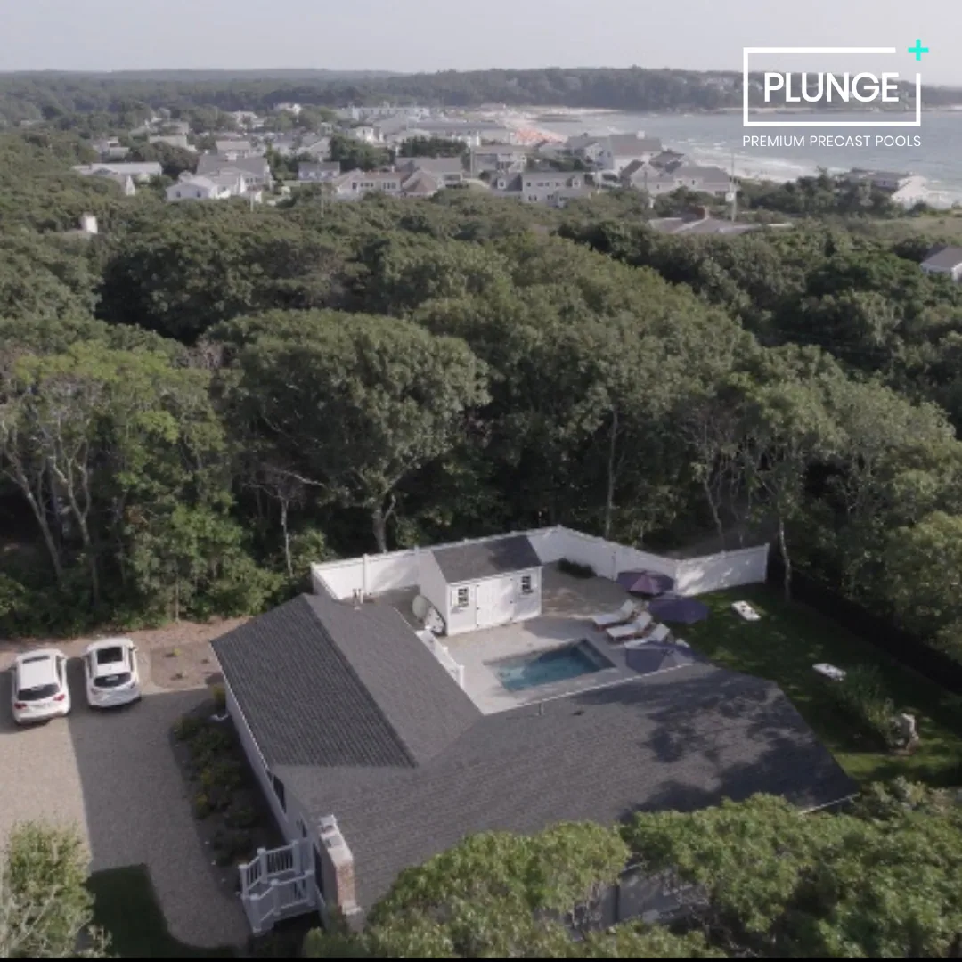 A house with a gray roof and a backyard featuring a pool and patio umbrellas, surrounded by trees and neighboring houses in the distance.
