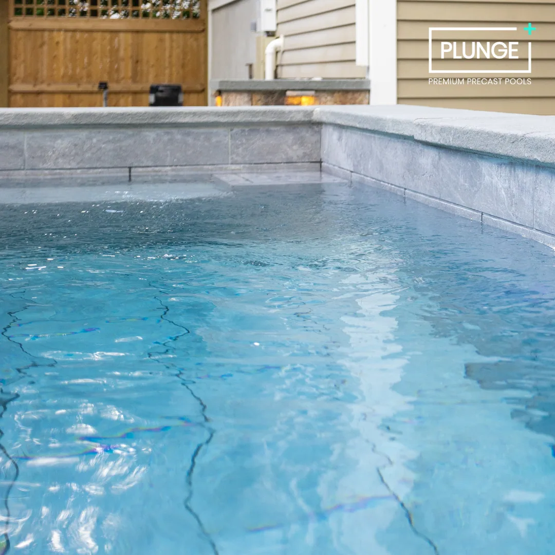 A close-up view of a luxury residential swimming pool with clear blue water, stone tile edges, in a backyard with a wooden fence and beige house siding in the background, featuring a logo that reads 'PLUNGE Premium Precast Pools'.